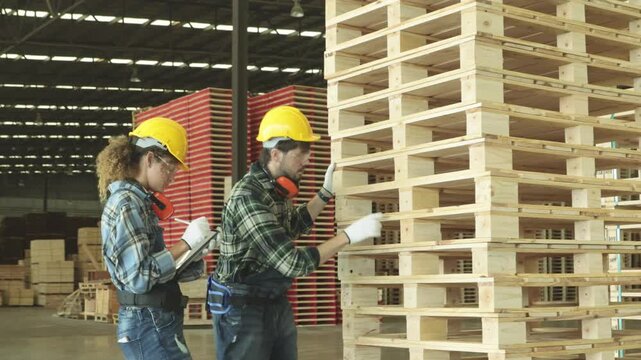 Male and female carpenter team wearing hard hats and safety glasses measure red painted pallet lumber tape measure the organized stacking handling these materials industrial facility classification.
