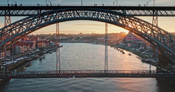 Drone flying in iconic Dom Luis I Bridge metal arch structure over the Douro River, with the historic Ribeira district of Porto city visible along the banks, bathed in the warm glow of sunset light