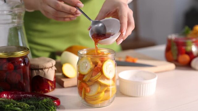 Making pickles. Woman adding sauce into glass jar with zucchini and pouring water at white wooden table indoors, closeup