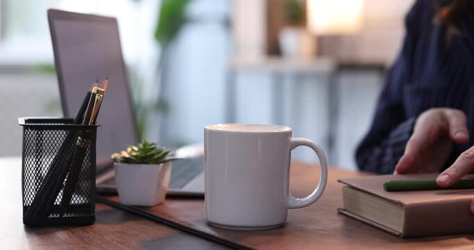 Woman putting mug of coffee and working with laptop at wooden table indoors, closeup