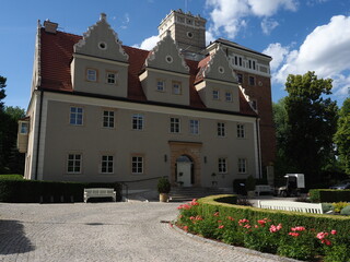 Topacz Castle in Poland with Decorative Gables, a Crenellated Tower and Red Tiled Roofs, Viewed across a Gravel Forecourt with Roses beneath a Blue Sky © Bluemoment72
