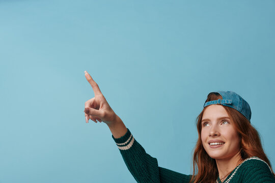 Smiling woman with red hair wearing a backward cap pointing upward against a blue background, casual fashion and confident mood, lifestyle image for wellness and imagination.