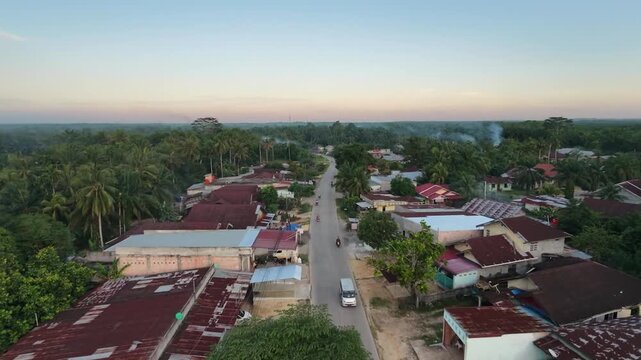 Aerial view of a rural village road passing through houses and lush palm oil plantations under a warm sunset sky. Tropical countryside community and evening atmosphere drone shot