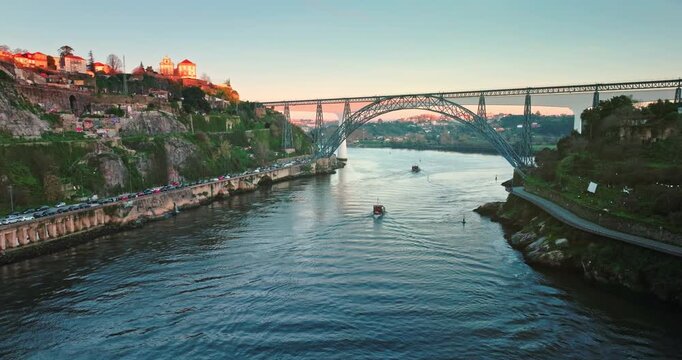 Aerial view traditional rabelo boats navigating the Douro River below the iconic Dom Luis I Bridge, connecting the historic cities of Porto and Vila Nova de Gaia at sunset. Travel and explore Portugal