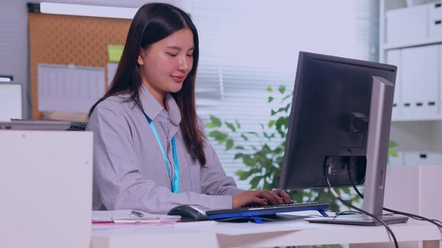 Asian woman office worker typing computer monitor organized desk modern workplace, combining focused professional expression, calm business atmosphere, writing activity, and productive office work