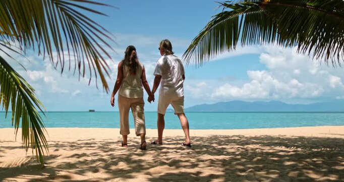 Senior couple holding hands while walking along sandy beach in Samui, Thailand, enjoying a tropical vacation with palm tree leaves framing the vibrant blue ocean and clear sky. Exotic travel getaway