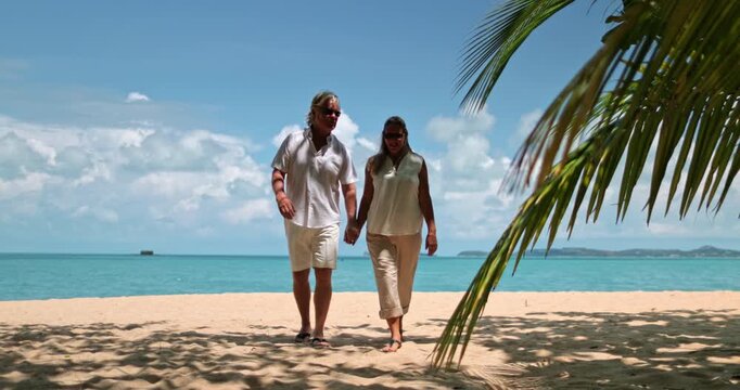 Happy senior couple holding hands, smiling, walking on a sandy beach under a palm tree, enjoying a romantic and relaxing holiday vacation at a resort on beautiful Koh Samui tropical Island, Thailand