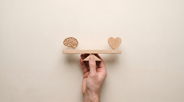 Hand balancing wooden icons of a brain and a heart on a seesaw scale, representing the delicate harmony between logic and emotion, rational thinking and passion in decision making and life.