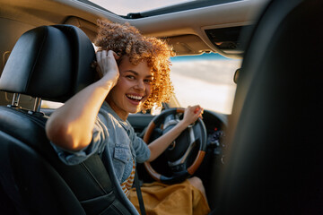 Obraz premium A young woman with curly hair smiles while driving a car in daylight, wearing a denim shirt and a seatbelt, capturing a bright casual moment on the road.
