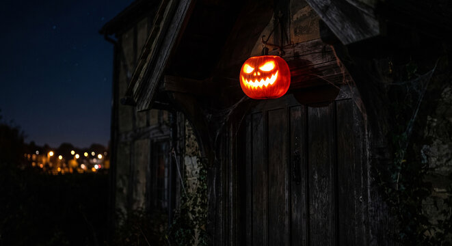 A Halloween pumpkin carved with a friendly face that appears to glow hangs in front of an old house.