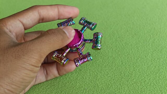 A hand holding a stationary rainbow metallic fidget spinner with hammer-shaped prongs on green background 