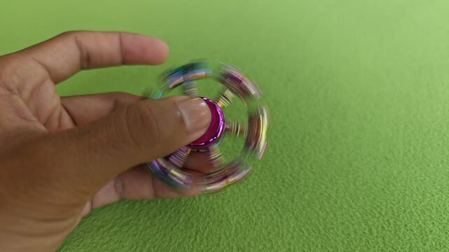 A hand rapidly spinning a colorful metallic hammer-shaped fidget spinner on green background