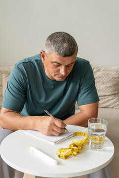 Man monitoring body measurements and weight at home. Using measuring tape, notebook, water, and an injection pen. Concept of metabolic health and healthy lifestyle
