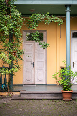 Fototapeta premium Weathered wooden door stands beneath green veranda roof beside ochre wall, framed by climbing vines, leafy shrubs, column, potted plant and paved courtyard with traditional residential architecture.
