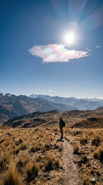 A lone tiny traveler standing in a vast high-altitude grassland in the Peruvian Andes, a small but vivid iridescent cloud glowing pastel pink, green and violet beside the sun, distant mountain ridges
