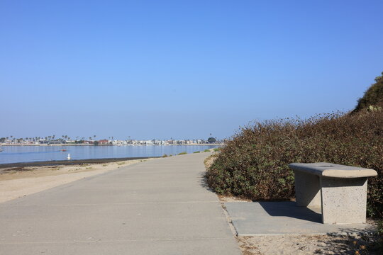 A quiet concrete path winds along Mission Bay with a stone bench nestled next to dry shrubs in San Diego.