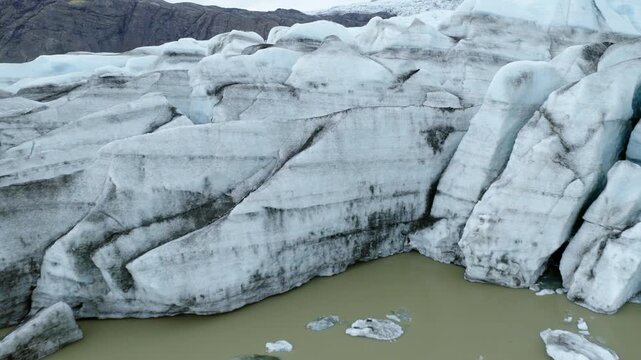 Fjallsjokull outlet glacier forms a heavily fractured terminus at Fjallsarlon in Orafi, South Iceland, with dark moraine bands and floating ice in the proglacial lagoon, drone pullback.