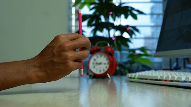 Hand holding pink pencil at home office desk, Close-up of a hand holding a pink pencil above a desk beside a computer keyboard, monitor, red alarm clock and leafy plant in a home office.