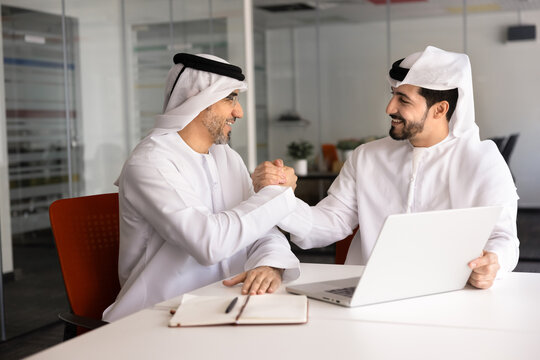 Smiling Middle-Eastern businessmen dressed in traditional Arab wear shaking hands sit at office desk with laptop, finalizing effective teamwork, business planning, professional discussion successfully