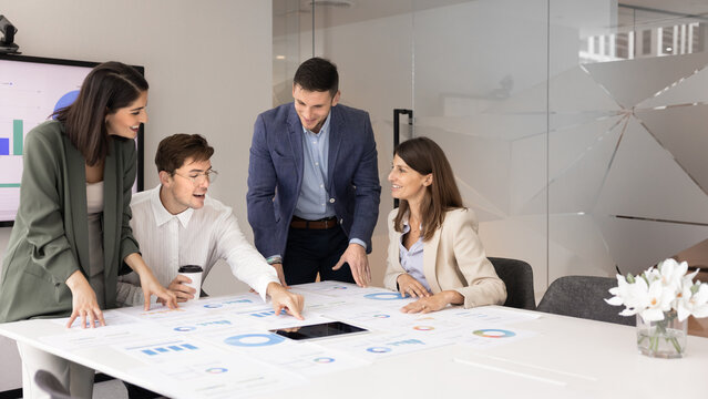 Friendly multicultural business professionals group teamworking on financial analytics in modern boardroom comparing digital and paper statistical reports on table setting company goals. Copy space