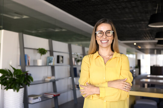 Portrait of young female office employee, corporate staff member stands with arms crossed, posing for professional photo at modern workplace, exuding success, leadership, and positivity. Career growth