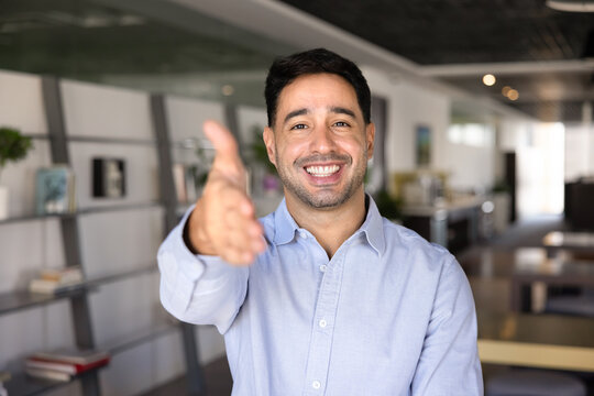 Head shot portrait of smiling businessman welcoming client, extending hand in greeting gesture to camera in office, starting negotiations or job interview. Partnership, collaboration, business deal