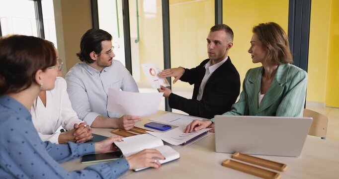 Young adult male executive presenting marketing report to diverse teammates on staff briefing at boardroom table analyzing results explaining charts and diagrams to colleagues during group evaluation