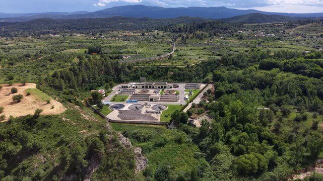 Water treatment plant surrounded by lush green forest and rolling hills, with circular clarifier tanks, industrial buildings, and a winding road visible in the background