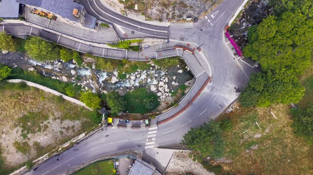 Top-down view of a winding mountain road intersection beside a rocky stream with parked cars, green vegetation and stone walls in a European alpine village