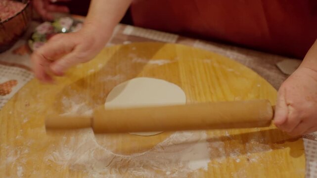 Cook flattens round dough piece with wooden rolling pin on circular cutting board while making authentic Georgian khinkali dumplings in Tbilisi kitchen.
