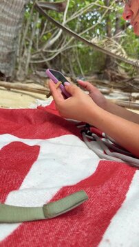 Side view of a woman tapping her smartphone while resting on a red and white beach towel beneath leafy trees. Sand, exposed roots and branches surround this quiet moment of outdoor holiday downtime.