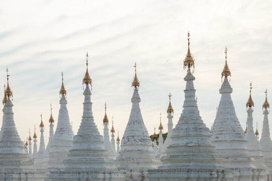 Rows of ornate white Buddhist stupas with golden finials at Kuthodaw Pagoda in Mandalay, Myanmar, creating a layered architectural pattern against an overcast sky