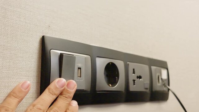 Hand turning on modern electrical wall switch next to power outlets and USB ports.