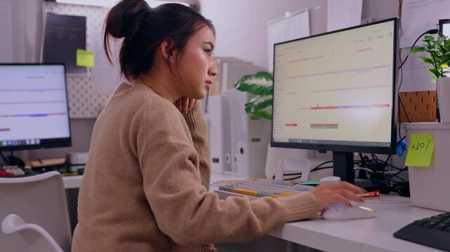 Female Asian office worker typing at computer monitor in modern workplace, focused expression, organized desk, managing digital workflow with calm concentration at workstation during remote work