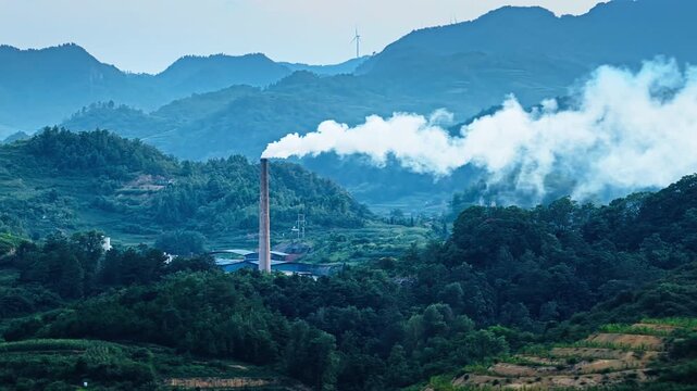 Industrial chimney spewing thick white smoke in a lush mountainous valley, highlighting environmental impact and pollution.