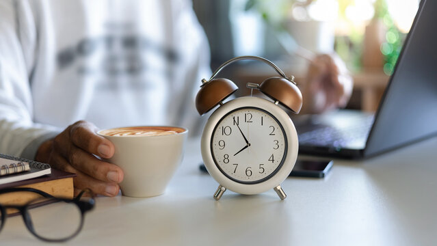 close up shot, alarm clock at 8 AM. blurred background with asian man using laptop working on desk and holding coffee cup. work or study in the morning after wake up