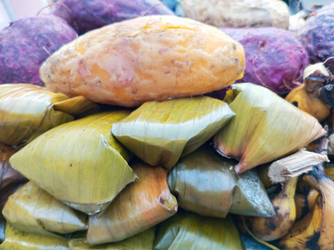 Close up view of traditional Indonesian steamed cake wrapped in banana leaves topped with a steamed sweet potato at a market counter