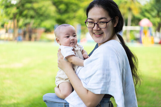 Mother and baby active lifestyle concept, smiling Asian mother wearing glasses carrying happy infant in hip seat carrier in green park, representing maternal care, parenting, and wellness.
