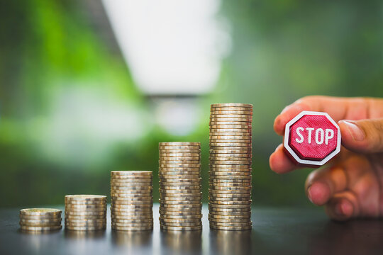 Financial limit and stop inflation concept, hand holding red stop sign next to tallest stack of silver coins on table, symbolizing capping expenses, budget ceiling, and interest pause.