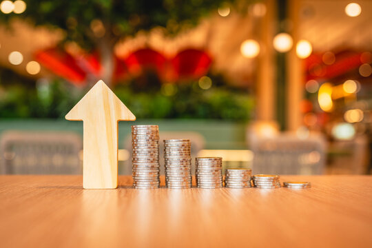 Upward economic trend and financial growth concept, wooden arrow pointing up standing beside ascending stacks of coins on table, symbolizing capital accumulation, profit, and revenue rise.