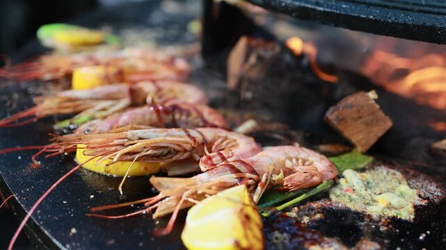 Shrimp grilling on round charcoal grill with lemon slices and garlic sauce. Barbecue seafood cooking over burning wood and coals, outdoor summer food preparation concept.