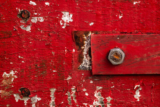 Close-up background of a weathered and peeling red painted wooden surface, featuring a rustic red metal bar attached with a rusty metallic bolt and old nails in Brazil.