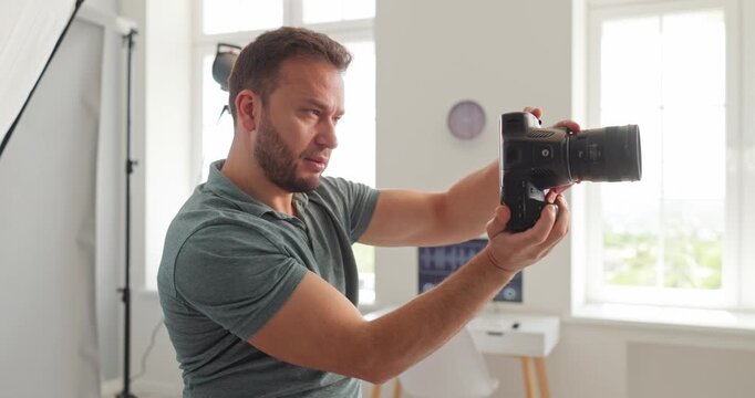 Photographer man focused with camera in studio. Focused professional checks settings for a portrait, adjusting angle near soft lighting equipment. Clear concept of precision and expertise.
