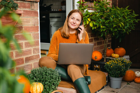 Woman talks on phone with laptop on autumn porch, working from home