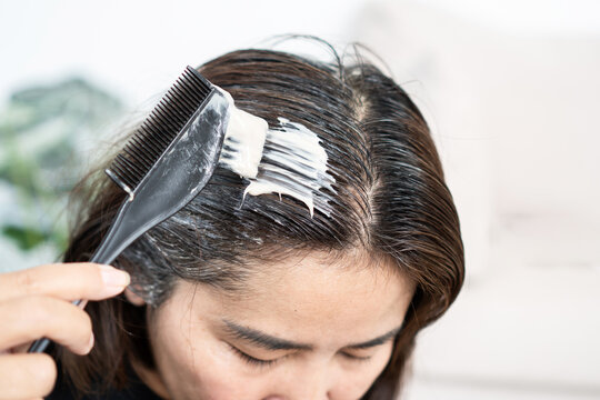 Middle-aged Asian woman applying dye cream on hair roots with brush to cover gray hair at home, haircare treatment, beauty salon coloring, personal grooming and aging wellness concept
