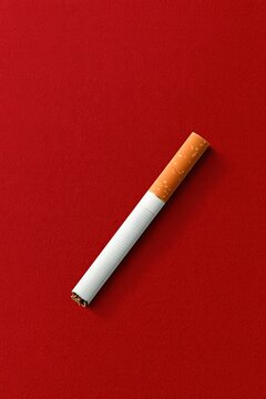 Single unlit cigarette with orange filter and white paper placed diagonally on solid deep red background showing smoking and tobacco product concept