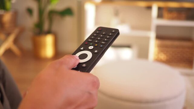 Close up of a person's hand pressing buttons on a remote control in a bright room.