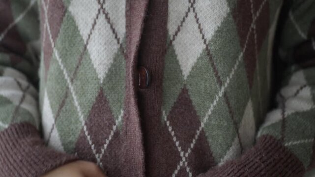 Close up of hands buttoning argyle pattern cardigan, slow motion, cozy knitwear fashion.