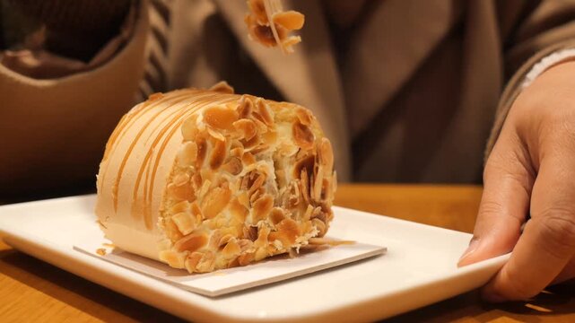 Person cutting into delicious almond roll cake using wooden fork on white plate in cafe.
