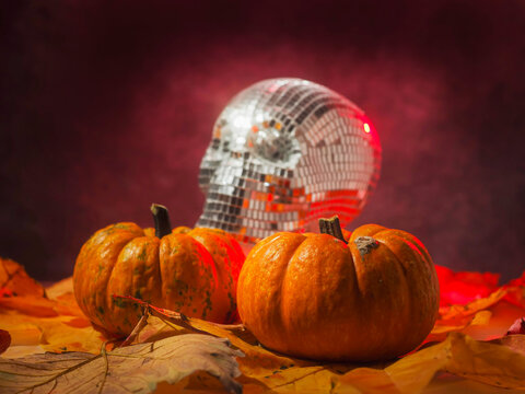 Disco ball skull glows beside mini pumpkins. Red smoke swirls around the dark scene. Autumn leaves scatter beneath the objects. No people present in the image. Perfect for Halloween or party themes